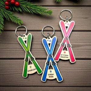 Peut inclure: Porte-clés en bois avec des skis croisés verts, bleus et roses. Chaque porte-clés a un anneau argenté et un nom de station de ski : Sugarloaf, Sunday River et Attitash Mountain. Les porte-clés sont sur une surface en bois avec une branche de verdure.