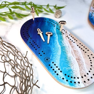 May include: A wooden cribbage board with a blue and white ocean wave design. The board has a compass in the center and is surrounded by holes for pegs. There are three wooden pegs with different designs on the board.