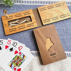 May include: A wooden cribbage board set with a brown leather case featuring a cutout of the state of Maine. The set includes wooden pegs and playing cards. The case is engraved with "MAINE - EST. 1820".