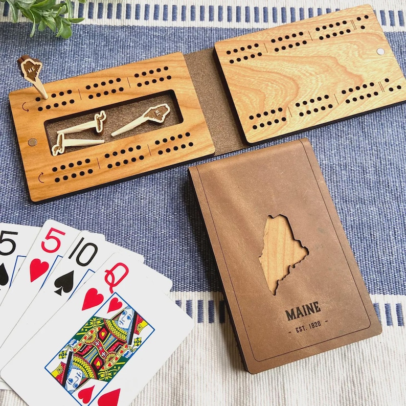 May include: A wooden cribbage board set with a brown leather case featuring a cutout of the state of Maine. The set includes wooden pegs and playing cards. The case is engraved with "MAINE - EST. 1820".