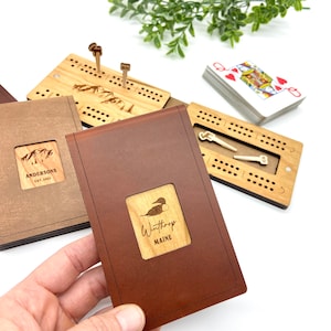 May include: A collection of wooden cribbage boards and a deck of cards. One board is in a brown leather case with a wooden inlay that reads "Winthrop Maine". Another board has a mountain design and the text "Andersons Est. 2021".