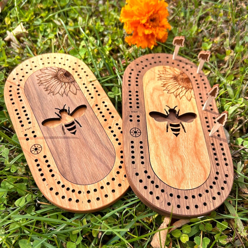 Peut inclure: Deux jeux de cribbage en bois avec un motif d'abeille et de tournesol. Les planches sont de forme ovale avec une bordure en bois fonc&eacute; et un centre en bois plus clair. Le centre pr&eacute;sente une abeille et un tournesol d&eacute;coup&eacute;s au laser. Des chevilles sont incluses.