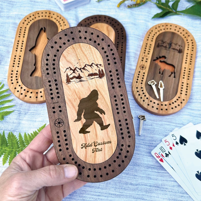 Peut inclure: Planches de cribbage en bois avec des motifs uniques. L'une d'elles pr&eacute;sente une silhouette de Bigfoot, des montagnes et le texte "Add Custom Text". D'autres planches montrent un loup, un &eacute;lan et un paysage. Chaque planche a une piste pour les chevilles et une rose des vents.