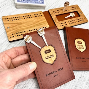 Peut inclure: Un jeu de société de cribbage en bois avec un étui en cuir marron. Il présente des motifs du "Parc national d'Acadia" et du "Grand Canyon National Park". Comprend des chevilles en bois et un jeu de cartes. Le design Acadia inclut le texte "1916".