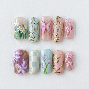 Puede incluir: Un conjunto de diez uñas postizas con varios diseños florales y de mariposas. Las uñas presentan una gama de colores, incluyendo rosa claro, azul y blanco, con detalles dorados. Los diseños incluyen margaritas, mariposas y patrones florales.