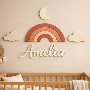May include: A wall-mounted decorative set featuring a layered rainbow in shades of terracotta and peach, a cream-coloured crescent moon, two cream-coloured clouds, and the name Amelia in cream-coloured script lettering mounted above a wooden cot.