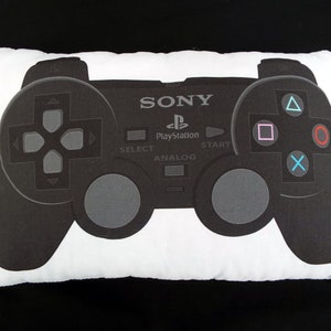 May include: A white rectangular pillow featuring a black and gray graphic of a PlayStation controller. The controller design includes the Sony logo, PlayStation branding, and button symbols in various colors. The pillow is a gaming-themed decorative item.