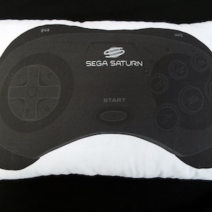 May include: White rectangular pillow featuring a black Sega Saturn controller design. The controller graphic includes the Sega Saturn logo, directional pad, buttons, and the word "START". A decorative item for gamers.