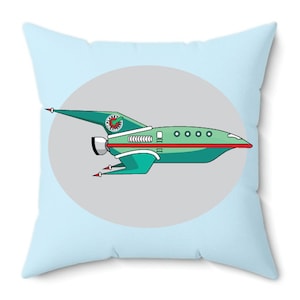 May include: A square throw pillow with a light blue background. The pillow features a cartoon spaceship design in shades of green, red, and white, set against a gray oval. The spaceship has a clock detail on the tail.