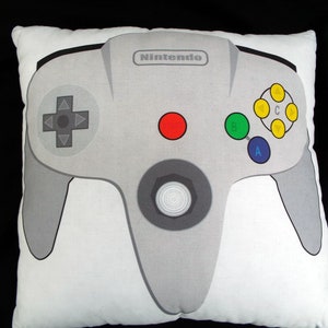 May include: Square white pillow featuring a gray Nintendo 64 controller design. The controller has a directional pad, colored buttons, and the Nintendo logo. A fun decorative item for gamers.