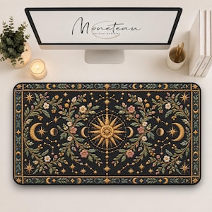 May include: A rectangular desk mat featuring a black background with a gold and multi-colored floral pattern, a central sun motif, and crescent moons. The design includes intricate botanical vines, small stars, and a decorative border.