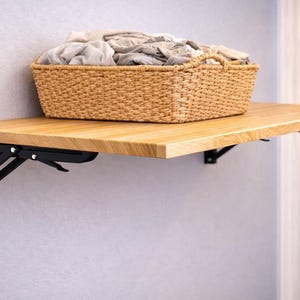 Op de afbeelding: Een lichtgekleurde houten opklapbare plank met zwarte metalen beugels, gemonteerd op een lichtgrijze muur. Een gevlochten mand gevuld met gevouwen lichtgekleurd linnen staat op de plank. De plank is ontworpen om op te vouwen wanneer deze niet in gebruik is.