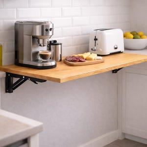 May include: A wall-mounted folding wooden table with black metal brackets, supporting a stainless steel espresso machine, a white toaster, and a wooden cutting board with sliced cheese and cured meats in a kitchen with white subway tile walls.