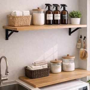 Op de afbeelding: Een wasruimte met houten planken met gevouwen witte handdoeken, glazen potten met houten deksels en bruine spuitflessen met de tekst "All Purpose" en "Cal Cleaner". Een wasmachine staat eronder.