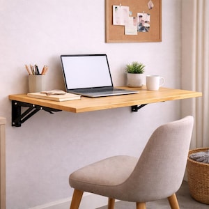 Peut inclure: Un bureau mural en bois clair avec un support en métal noir. Un ordinateur portable, une tasse blanche et une petite plante en pot sont posés sur le bureau. Une chaise beige et un panier en osier sont devant le bureau.