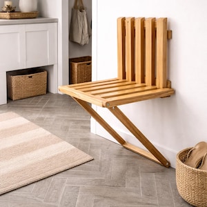 Könnte beinhalten: Wandmontierter Klappstuhl aus hellem Holz. Der Stuhl hat eine Sitzfläche und Rückenlehne aus Latten, mit abgewinkelten Stützbeinen. Der Hintergrund zeigt einen Teppich und einen Korb. Maße: ca. 40 cm x 30 cm.