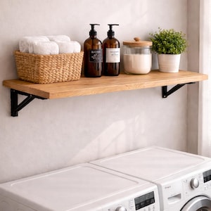 Op de afbeelding: Een houten plank met zwarte beugels houdt een geweven mand met witte handdoeken, twee bruine pompflessen, een glazen pot met een houten deksel en een plant in een pot. Daaronder zijn twee witte wasmachines zichtbaar.
