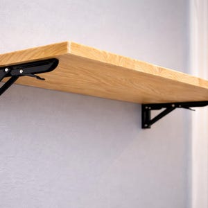 Op de afbeelding: Een lichtgekleurde houten plank die aan de muur is bevestigd met zwarte metalen opvouwbare beugels. De plank is rechthoekig en lijkt van massief hout te zijn. De beugels zijn aan de muur bevestigd en ondersteunen de plank, waardoor deze kan worden opgevouwen wanneer deze niet in gebruik is.