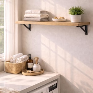 Op de afbeelding: Een wasruimte met een houten plank met opgevouwen handdoeken, een kleine plant in een pot en een witte kom met decoratieve items. Daaronder een wasmachine, een gevlochten mand en een dienblad met toiletartikelen.