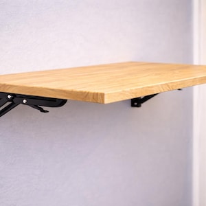以下が含まれることがあります： 明るい色の木製壁掛け折りたたみテーブル。 テーブルは黒い金属製の折りたたみブラケットで支えられています。 テーブルは長方形で、無垢材でできているようです。 ブラケットは薄い灰色の壁に取り付けられています。