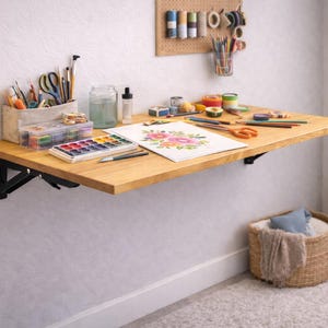 Peut inclure: Un bureau mural en bois clair avec des supports en métal noir, contenant du matériel d'art. Une palette d'aquarelle, des crayons, des ciseaux et un dessin de fleurs sont sur le bureau. Un panier avec une couverture est en dessous.