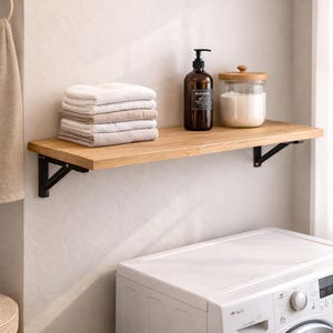 May include: A wooden shelf with black metal brackets holds a stack of folded towels, a brown soap dispenser, and a glass jar with a wooden lid. The shelf is mounted above a white washing machine in a laundry room.