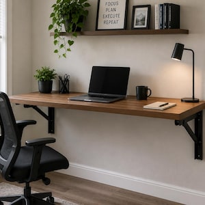 Könnte beinhalten: Ein wandmontierter Schreibtisch aus Holz mit schwarzen Metallhalterungen, ausgestattet mit einem Laptop, einer schwarzen Tasse, einem Notizbuch mit Stift und einer schwarzen Schreibtischlampe. Darüber befindet sich ein Regal mit Pflanze und Schild.