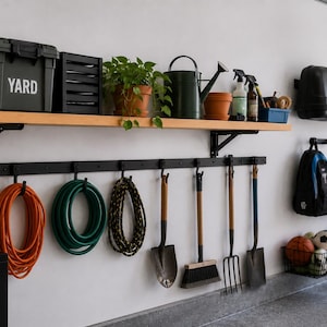 May include: Garage organization system with a wooden shelf, black storage bin labeled "YARD", and hanging tools. Includes an orange hose, green hose, and various gardening tools. A black toolbox and two bicycles are also visible.