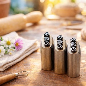 May include: Three metal stamps with Chinese characters are displayed on a wooden surface. A small bouquet of purple and white flowers rests on a folded cloth. A rolling pin and pottery tools are also visible.