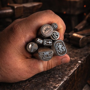 Könnte beinhalten: Nahaufnahme einer Hand, die eine Sammlung von Metallstempeln mit verschiedenen Designs hält, darunter Hammer, Amboss und Schild. Die Stempel sind grau mit schwarzen Details. Im Hintergrund eine Schmiede.