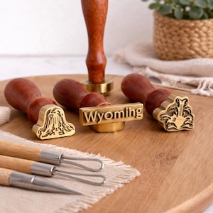 Puede incluir: Una colección de sellos con mango de madera y cabezales de sello de latón, incluido uno que dice "Wyoming". Los sellos están dispuestos sobre una tabla de cortar de madera con un pequeño paño de lino y un juego de herramientas de metal.