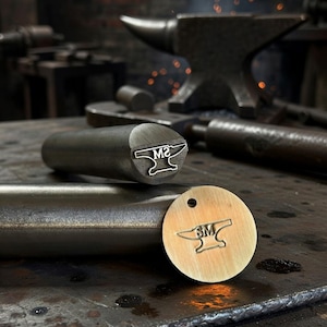 May include: Close-up of a metal stamp with the letters "M2" and an anvil design. A round brass tag with the same design is also visible. The background shows an anvil and other blacksmithing tools, suggesting a workshop setting.