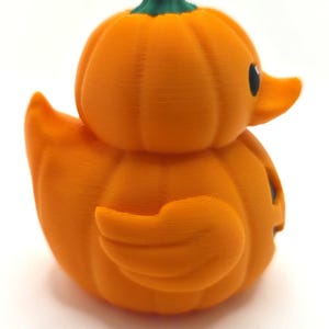 May include: A 3D printed rubber duck shaped like a pumpkin with a bright orange body, a green stem on top, and a black eye. The surface features visible horizontal layer lines from the printing process, creating a textured appearance.