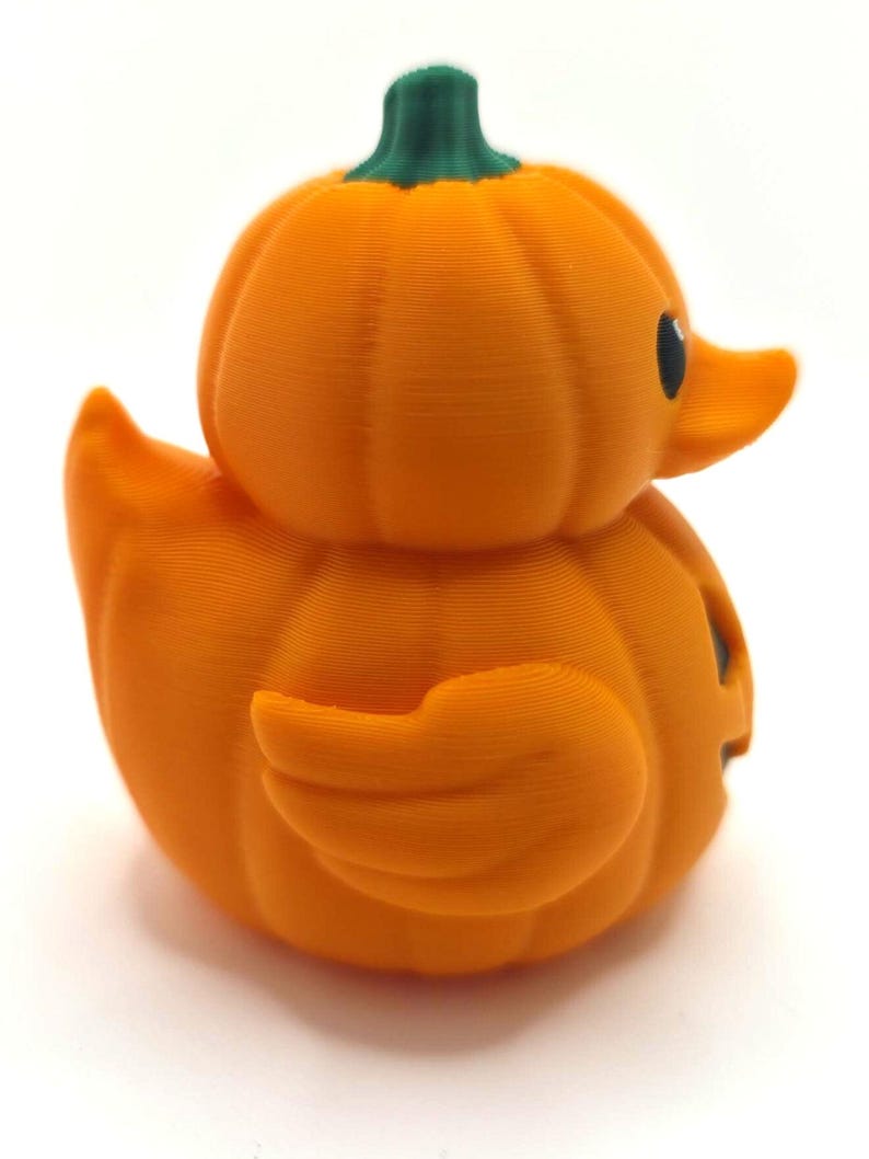 May include: A 3D printed rubber duck shaped like a pumpkin with a bright orange body, a green stem on top, and a black eye. The surface features visible horizontal layer lines from the printing process, creating a textured appearance.