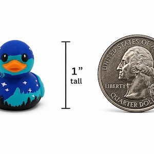 May include: A small blue rubber duck featuring a night sky design with white stars and a black forest silhouette at the base. The duck has an orange beak and is shown next to a United States quarter dollar coin, indicating a height of 1 inch.