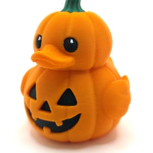 May include: A 3D printed rubber duck shaped like a pumpkin with a green stem on top. The duck has a bright orange body with a carved jack-o'-lantern face featuring black triangular eyes and a black mouth with two teeth on the front of its chest.