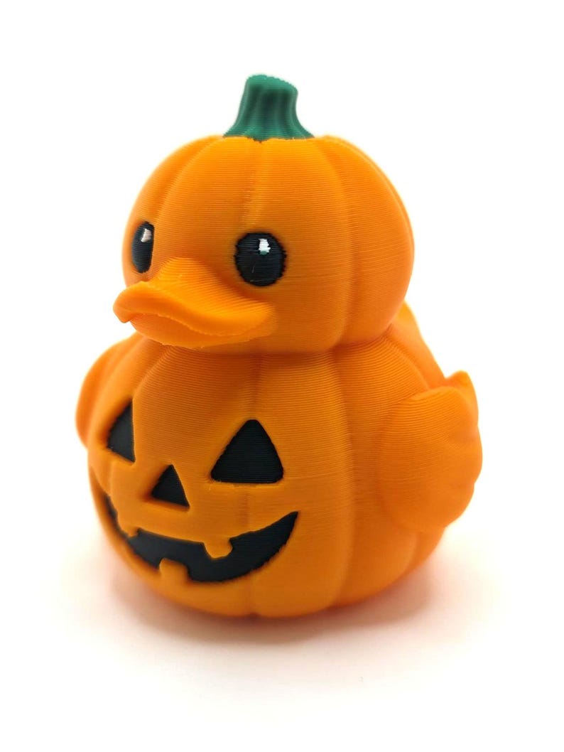 May include: A 3D printed rubber duck shaped like a pumpkin with a green stem on top. The duck has a bright orange body with a carved jack-o'-lantern face featuring black triangular eyes and a black mouth with two teeth on the front of its chest.