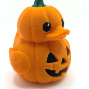 May include: A small orange rubber duck shaped like a pumpkin with a green stem on top. The duck features a carved jack-o'-lantern face with black triangular eyes and a black mouth. The surface has a visible 3D printed texture throughout.