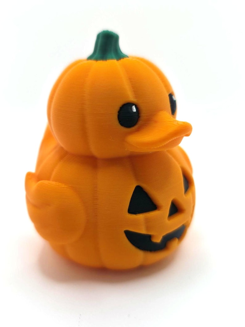 May include: A small orange rubber duck shaped like a pumpkin with a green stem on top. The duck features a carved jack-o'-lantern face with black triangular eyes and a black mouth. The surface has a visible 3D printed texture throughout.