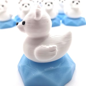 May include: A small white 3D printed polar bear figurine with black eyes and nose, perched atop a light blue hexagonal base. The surface of the bear shows horizontal layer lines from the printing process, and several identical figures are visible behind.