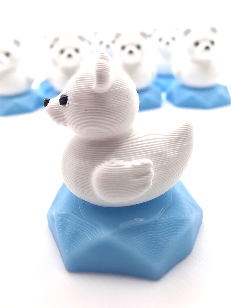 May include: A small white 3D printed polar bear figurine with black eyes and nose, perched atop a light blue hexagonal base. The surface of the bear shows horizontal layer lines from the printing process, and several identical figures are visible behind.