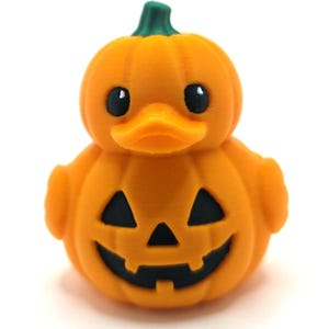 May include: A 3D printed orange rubber duck shaped like a pumpkin. The duck has a green stem on top, black eyes, and a carved jack-o'-lantern face on its chest with triangular eyes and a jagged mouth. The surface shows fine horizontal print lines.