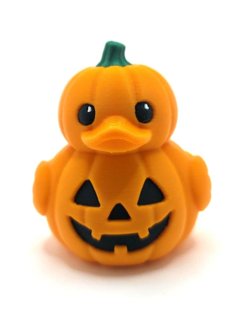 May include: A 3D printed orange rubber duck shaped like a pumpkin. The duck has a green stem on top, black eyes, and a carved jack-o'-lantern face on its chest with triangular eyes and a jagged mouth. The surface shows fine horizontal print lines.