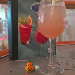May include: A stemmed glass filled with a pale pink cocktail, garnished with a lime wedge and a white straw, sits beside a small orange pumpkin-shaped rubber duck and a menu featuring illustrations of various colorful cocktails.