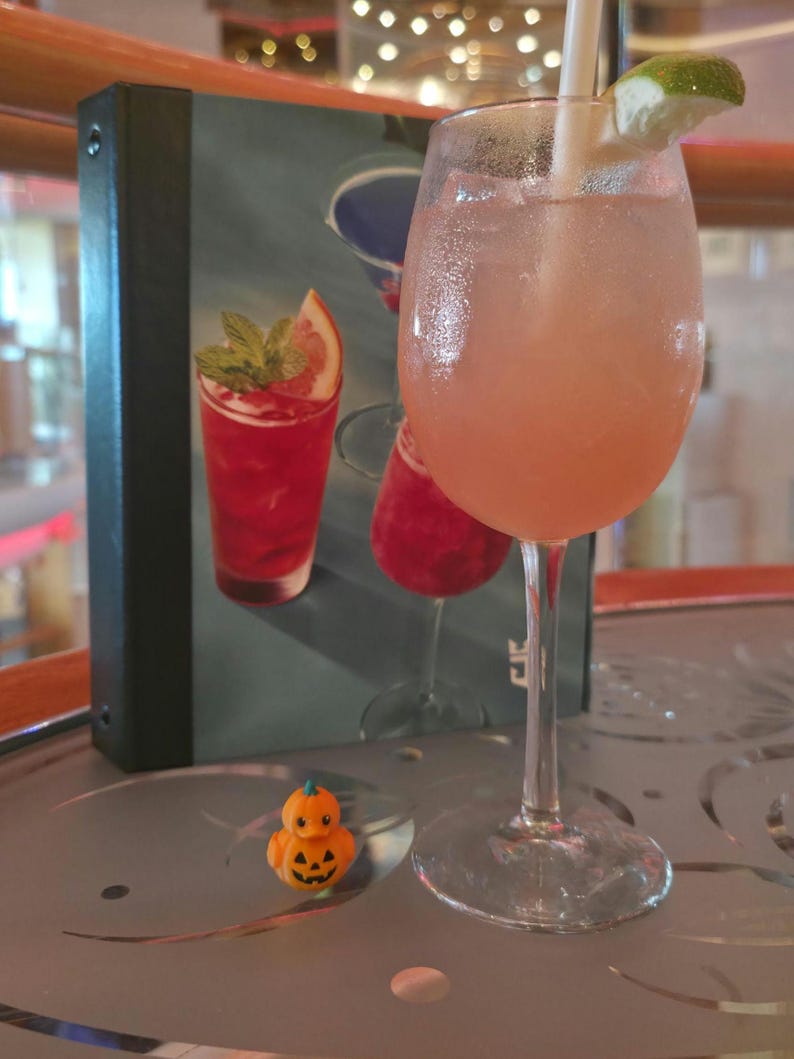 May include: A stemmed glass filled with a pale pink cocktail, garnished with a lime wedge and a white straw, sits beside a small orange pumpkin-shaped rubber duck and a menu featuring illustrations of various colorful cocktails.