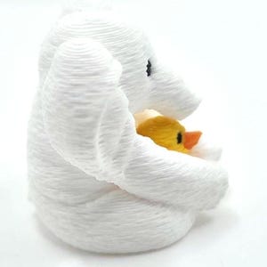 May include: A white 3D printed elephant figurine with a textured surface, featuring a small yellow duckling with an orange beak nestled within its trunk and front legs. The elephant has a black eye and a rounded, layered appearance.