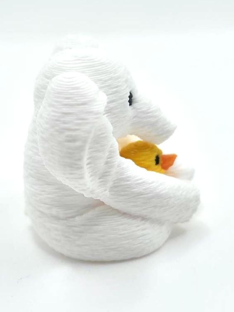 May include: A white 3D printed elephant figurine with a textured surface, featuring a small yellow duckling with an orange beak nestled within its trunk and front legs. The elephant has a black eye and a rounded, layered appearance.