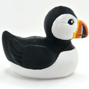 May include: A puffin figurine with a black head and back, white chest, and a large, colorful beak. The beak is orange, yellow, and black. The bird is sitting on a white surface with a blurred ocean background. 3d Printed in PLA Plastic.