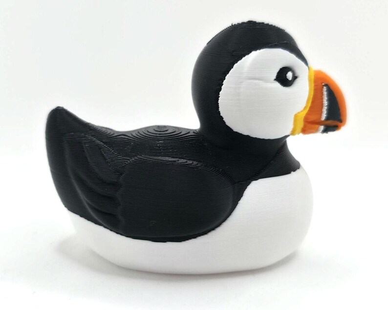 May include: A puffin figurine with a black head and back, white chest, and a large, colorful beak. The beak is orange, yellow, and black. The bird is sitting on a white surface with a blurred ocean background. 3d Printed in PLA Plastic.
