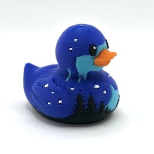May include: A 3D printed rubber duck figure featuring a dark blue body with a light blue face and an orange beak. The design includes a black silhouette of a forest at the base, white star-like dots across the body, and visible horizontal layers.